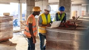 sécurité chantier - 3 hommes avec des casques de chantier discutant dans un immeuble en cours de construction avec un PC